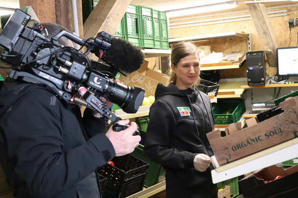 Eine Frau in schwarzer Arbeitskleidung hält eine Holzkiste mit der Aufschrift ‚Organic Soul‘, daneben ein Kameramann, der sie filmt. Im Hintergrund grüne Gemüsekisten.