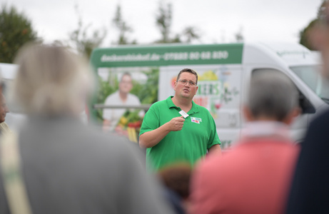 Ein Mann in einem grünen Hemd spricht mit einer kleinen Menschenmenge im Freien. Er hält ein Mikrofon und scheint eine Präsentation oder Ansprache zu halten. Im Hintergrund steht ein weißer Lieferwagen mit einem Werbeaufdruck für frische Lebensmittel, auf dem ebenfalls ein Bild des Mannes zu sehen ist. Einige Personen sind unscharf im Vordergrund zu erkennen und hören zu.