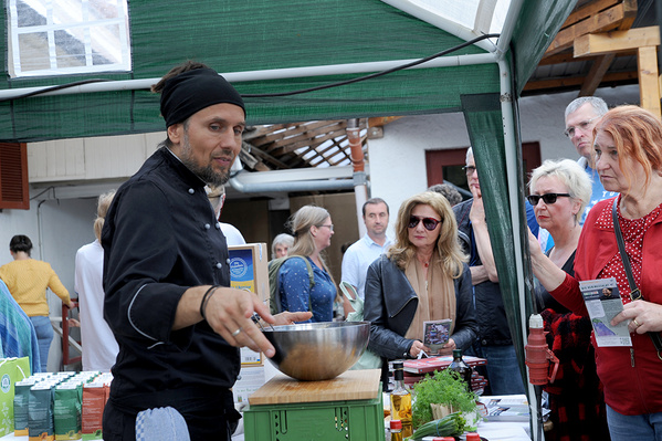 Ein Mann mit Bart und Kochmütze steht an einem Stand und hält eine Schüssel mit Essen in der Hand, während er mit den Besuchern spricht
