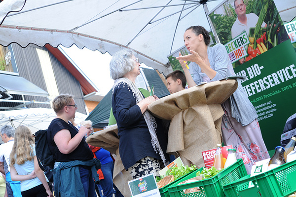 Zwei Frauen stehen an einem Stand auf einem Hoffest, der mit grünen Kisten gefüllt ist. Sie unterhalten sich mit einer Besucherin. Auf einem Banner ist die Aufschrift "BIO-Lieferservice" zu lesen.