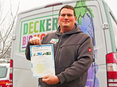  Ein Mann steht vor einem weißen Transporter und hält eine Urkunde in die Kamera. Er trägt eine graue Kapuzenjacke über einem hellgrünen Hemd und eine Brille. Auf dem Transporter ist in großen Buchstaben das Wort "DECKERS BIOHOF" und eine Abbildung einer großen violetten Aubergine zu sehen.