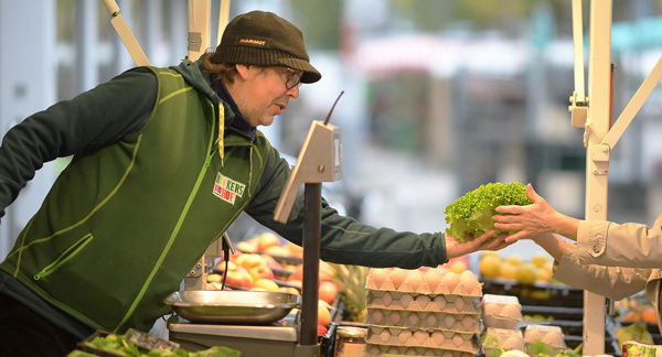 KI generiert: Ein Verkäufer reicht einem Kunden einen Salatkopf über den Tresen eines Marktstandes. Der Stand bietet frische Produkte wie Eier und Gemüse an.