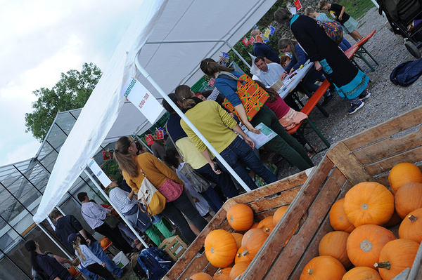 Eine Gruppe von Menschen sitzt und steht unter einem großen weißen Zelt an Tischen. Im Vordergrund sind Kisten mit Kürbissen gestapelt.