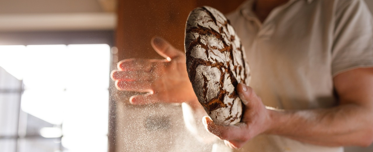  Ein Bäcker in heller Kleidung hält ein frisch gebackenes, rustikales Brot mit dunkler, rissiger Kruste in seinen Händen. Er klopft oder reibt das Brot ab, wodurch eine Wolke aus Mehlstaub oder Puderzucker in die Luft steigt. Das Licht fällt von links in den Raum und beleuchtet den Staub und die Hände des Bäckers.
