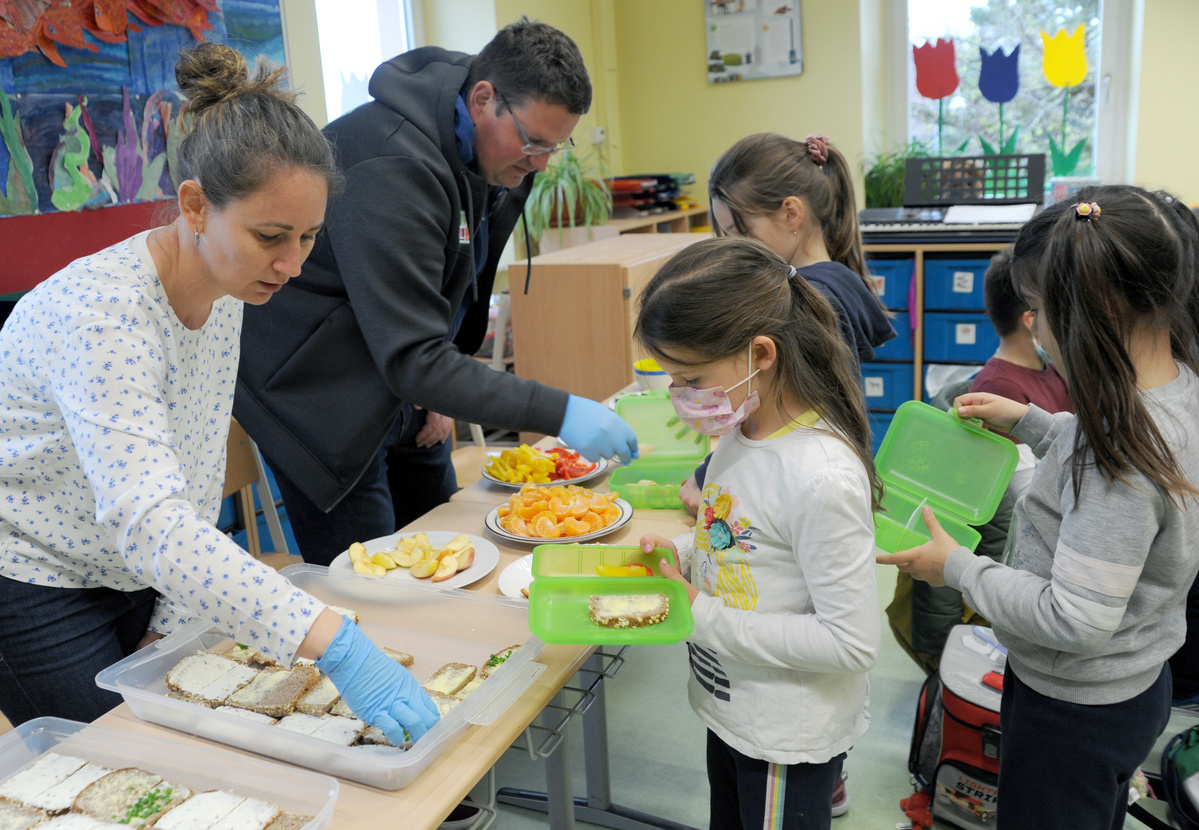 Zwei Erwachsene – eine Frau mit blau-weißer Bluse und ein Mann in einem grauen Hoodie – stehen hinter einem Tisch in einem Klassenraum und servieren Essen für eine Gruppe von Schulkindern. Die Frau verteilt belegte Brote aus einer Plastikbox, während der Mann Obstscheiben auf die Teller der Kinder legt. Ein Mädchen mit Gesichtsmaske hält eine grüne Brotdose offen, um sie füllen zu lassen.