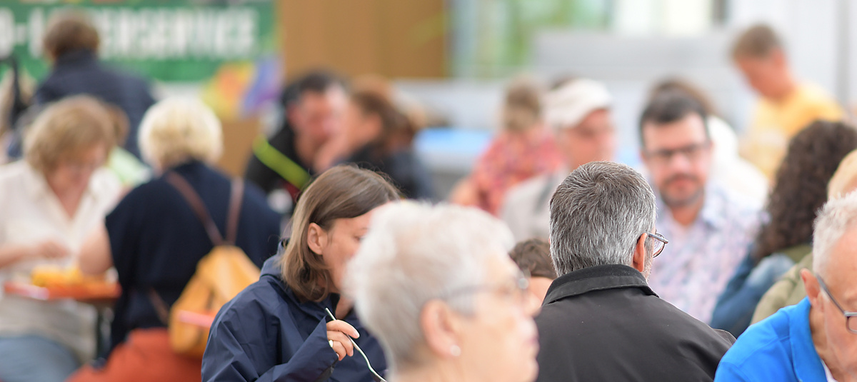 Viele Menschen sitzen dicht beieinander an langen Tischen im Freien. Es herrscht eine lebhafte Atmosphäre, mehrere Personen unterhalten sich und essen gemeinsam. Im Hintergrund sind unscharf weitere Leute und grüne sowie hölzerne Elemente zu erkennen. Die Szene wirkt wie ein Fest oder eine gemeinschaftliche Veranstaltung mit entspannter Stimmung. Manche tragen Jacken und Rucksäcke, einige haben graues Haar. Im Vordergrund hält eine Person Besteck in der Hand.