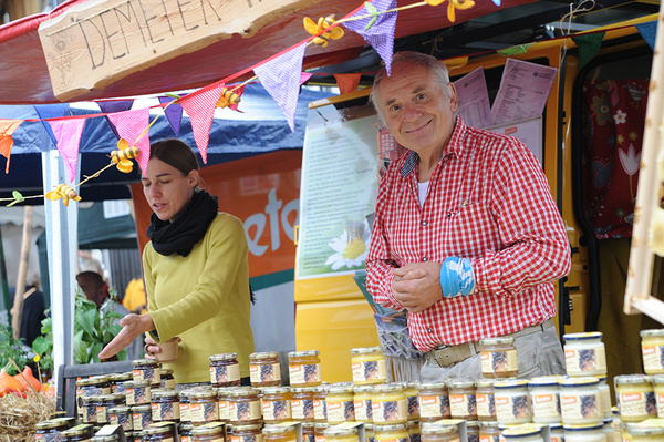 Ein Mann mit einem karierten Hemd lächelt an einem Marktstand, der mit bunten Wimpeln und einem Schild mit der Aufschrift "DEMETER" geschmückt ist. Daneben stehen Honiggläser, und eine Frau bedient Besucher.