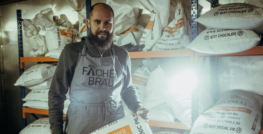  Ein bärtiger Mann mit einem grauen Pullover und einer hellgrauen Schürze mit der Aufschrift „FÄCHER BRÄU“ hält einen großen weißen Sack mit der Aufschrift „ORGANIC PILSNE“ in einem Lagerraum. Er steht zwischen Regalen, die mit weiteren weißen Säcken voller Malz, teilweise mit der Aufschrift „BESTMALZ“, gefüllt sind. Ein helles Licht strahlt von rechts unten ins Bild.