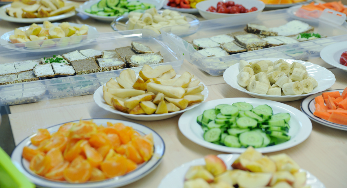 Eine Nahaufnahme eines gedeckten Tisches mit verschiedenen geschnittenen Lebensmitteln, die für ein Buffet vorbereitet sind. Es gibt flache Teller mit in Scheiben geschnittenen Birnen, Bananen, Gurken und Karottenwürfeln. Daneben stehen große Plastikboxen, die mit belegten Brotscheiben gefüllt sind.