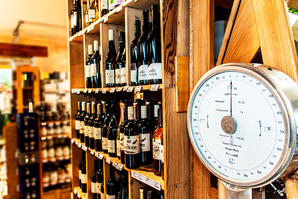 Eine Panoramaaufnahme, die eine Ladenszene mit Wein- und Käseprodukten zeigt. Auf der linken Seite sind Weinflaschen in Regalen zu sehen, die in der Mitte des Bildes scharf abgebildet sind. Auf der rechten Seite ist eine große, runde Waage mit einer Skala in Kilogramm zu sehen. Die Nadel zeigt genau auf 0 kg.