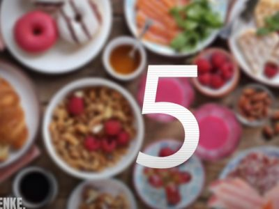 Dieses Bild zeigt einen unscharfen, von oben aufgenommenen Blick auf einen Tisch mit verschiedenen Gerichten. Man kann Donuts, Croissants, Müsli mit Himbeeren, Wurst, Käse, Honig und Nüsse erkennen. Eine große, weiße Zahl 5 steht im Vordergrund.