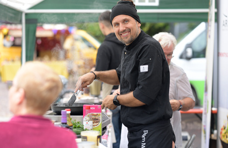 Ein Koch in schwarzer Jacke lächelt freundlich an einem Food-Stand mit regionalen Produkten – Teil der kulinarischen Verpflegung beim Fest.
