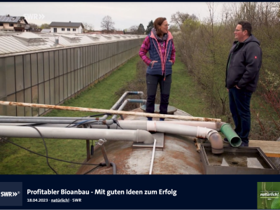 Screenshot einer Fernsehsendung des SWR mit dem Titel: Profitabler Bioanbau – Mit guten Ideen zum Erfolg. Zwei Personen stehen auf einem großen Wassertank neben einem Gewächshaus und unterhalten sich.