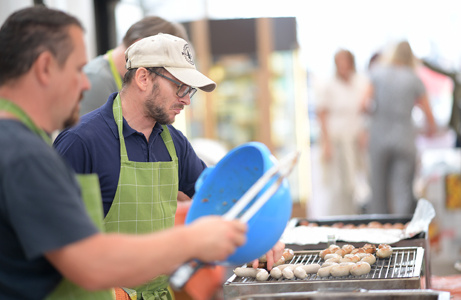 Zwei Männer grillen Essen im Freien, der eine hält eine Zange und der andere eine blaue Schüssel.