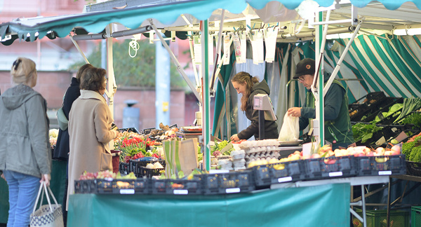 KI generiert: Das Bild zeigt einen Marktstand mit frischem Obst und Gemüse, an dem mehrere Personen einkaufen. Die Verkäufer bedienen die Kunden an einem offenen Stand mit Markise.
