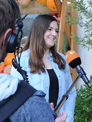 Eine junge Frau mit langen braunen Haaren in einem hellen Jeanshemd und einem schwarzen Top lächelt, während sie von einem Interviewer befragt wird, der nur teilweise im Bild zu sehen ist. Vor ihr ist ein Mikrofon mit einem orangenen Aufsatz, der ein Logo mit der Aufschrift "SWR" trägt, und Kopfhörer zu erkennen. Im Hintergrund sind Kürbisse und herbstliche Dekorationen zu sehen.