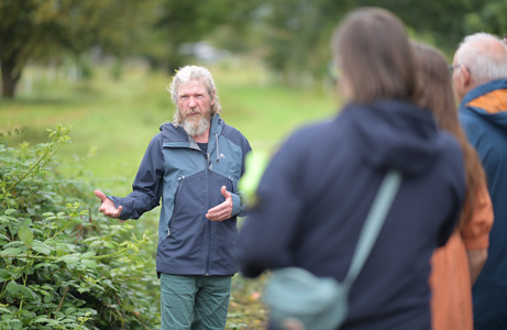 Ein Mann spricht gestikulierend mit einer Gruppe von Menschen im Grünen – bei einer Kräuterführung auf dem Feld.