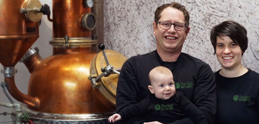 Ein lächelndes Paar steht mit einem Baby im Arm vor einer großen, glänzenden Kupferbrennblase. Der Mann trägt eine Brille und ein dunkles T-Shirt mit einem runden grünen Logo. Die Frau hat kurze dunkle Haare und trägt ebenfalls ein dunkles T-Shirt mit dem gleichen Logo. Das Baby trägt einen passenden dunklen Strampler mit dem Logo und blickt zur Seite.