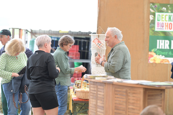 Eine Gruppe älterer Menschen steht an einem Honig-Verkaufsstand. Ein Mann in einem blauen Hemd und eine Frau mit blonden Haaren sind im Gespräch mit zwei anderen Personen.