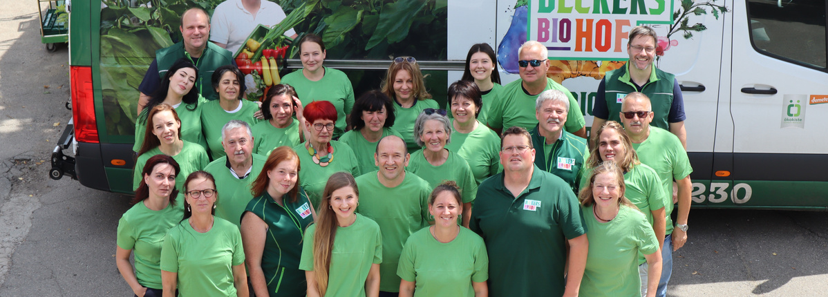 Gruppenfoto von Mitarbeitenden eines Biobetriebs vor einem Lieferwagen mit Aufschrift 'Biohof' und buntem Gemüse-Motiv. Die Personen tragen grüne Oberteile und lächeln in die Kamera