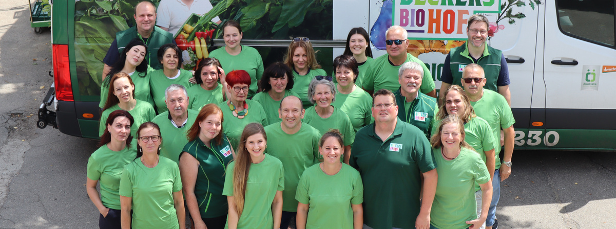 Gruppenfoto von Mitarbeitenden eines Biobetriebs vor einem Lieferwagen mit Aufschrift 'Biohof' und buntem Gemüse-Motiv. Die Personen tragen grüne Oberteile und lächeln in die Kamera