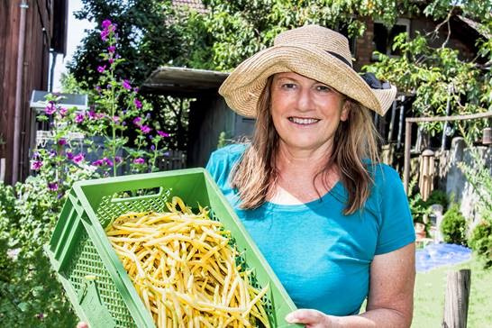Helga Decker mit einem großen Strohhut und einem blauen T-Shirt lächelt in einem Garten oder auf einem Feld direkt in die Kamera. Sie hält eine grüne Kiste mit gelben Paprika in der Hand. Im Hintergrund sind blühende lila Blumen und grünes Laub zu sehen.