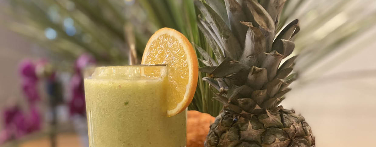 Eine Nahaufnahme von einem frisch zubereiteten, gelb-grünen Smoothie in einem Glas, das mit einer Orangenscheibe am Rand dekoriert ist. Im Hintergrund sind unscharf eine Ananas und andere Früchte sowie Pflanzen zu sehen, was auf ein tropisches Ambiente hindeutet.
