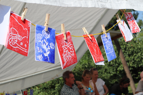 Mehrere bunt bemalte und verzierte Papiere hängen an einer Leine unter einem Zeltdach. Im Hintergrund sind undeutlich sitzende Personen zu erkennen.