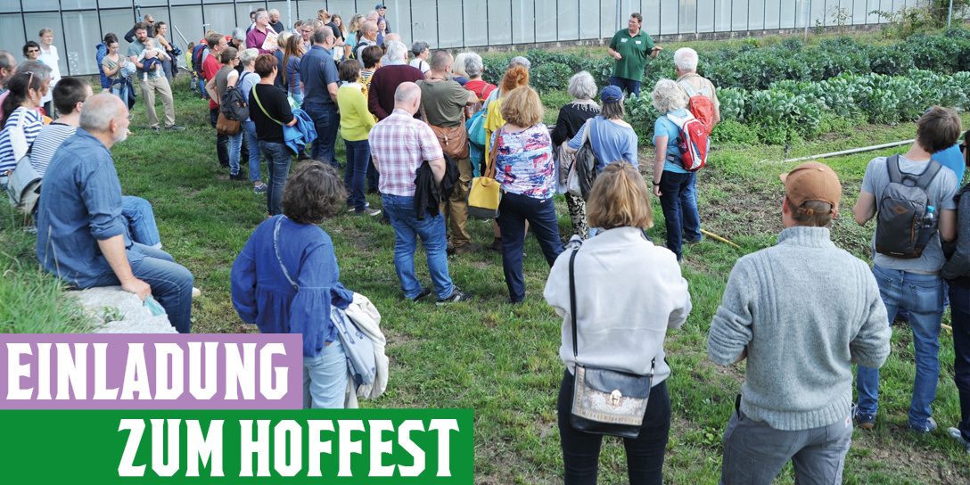 Eine Gruppe von Menschen steht auf einem Bauernhof im Grünen und hört einem Mann zu, der vor einem Gemüsebeet spricht. Im Vordergrund ein Schild mit der Aufschrift „Einladung zum Hoffest“. Die Besucher:innen sind unterschiedlichen Alters und aufmerksam der Führung zugewandt.