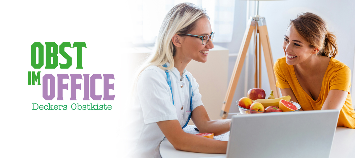 Zwei Frauen sitzen gemeinsam vor einem Laptop in einem Büro. Eine Frau in weißem Oberteil erklärt etwas, die andere in einem orangen Shirt hört zu. Im Vordergrund steht ein Obstkorb mit Äpfeln und Bananen.