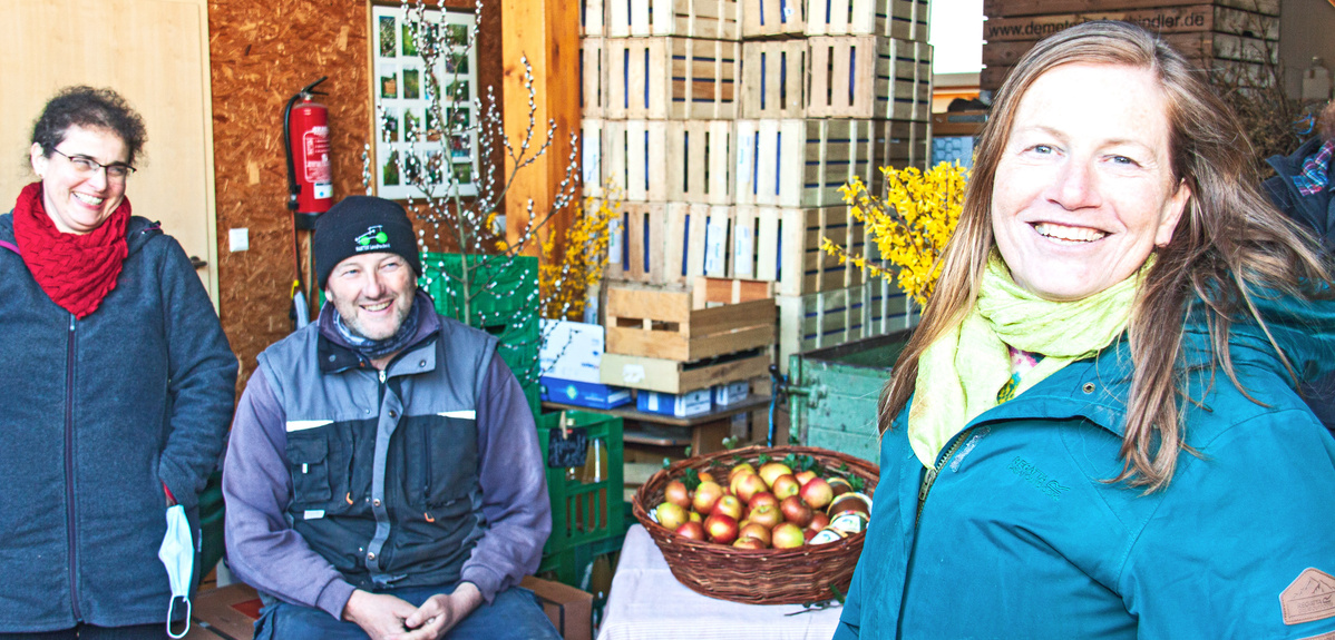  Drei lächelnde Personen stehen oder sitzen in einem Lagerraum, der zur Kontrolle, Sortierung und Lagerung von Äpfeln dient. Im Hintergrund sind gestapelte Holzkisten und ein Feuerlöscher zu sehen. Im Vordergrund rechts lächelt eine Frau mit langen Haaren in einer grünen Jacke und einem hellgrünen Schal direkt in die Kamera. Neben ihr sitzt ein Mann mit Mütze und Bart, der ebenfalls lächelt. Links steht eine Frau mit lockigen Haaren und einem roten Schal, die auch lächelt. Vor dem Mann und der Frau rechts steht ein Weidenkorb voller Äpfel.