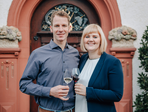Weinbauernpaar steht mit Weingläsern vor rotem Gebäude.