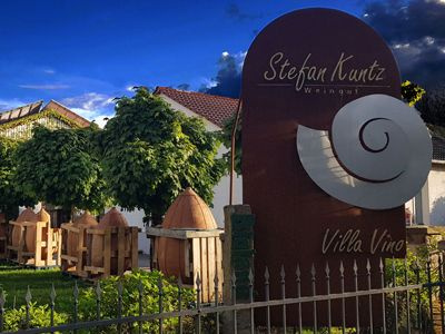 Schild mit der Aufschrift ‚Stefan Kuntz Villa Vino‘ vor Weinreben und Haus.
