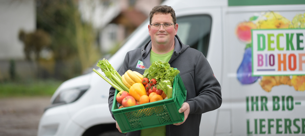 Christoph Decker steht vor einem Lieferwagen und hält eine grüne Kiste mit frischem Bio-Obst und -Gemüse, darunter Bananen, Äpfel, Tomaten, Paprika und Sellerie. Auf dem Lieferwagen steht das Logo ‚Deckers Biohof‘.