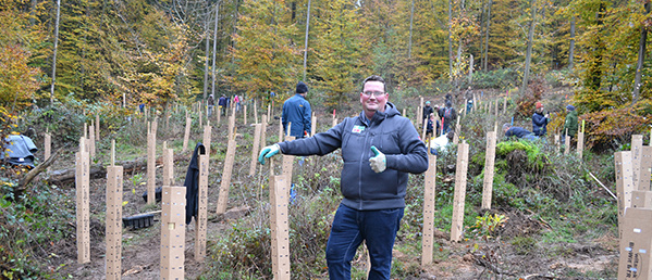 Ein Mann steht lächelnd in einem lichten Waldstück und gibt den Daumen hoch. Er trägt eine graue Jacke, eine Brille und grüne Arbeitshandschuhe. Um ihn herum sind zahlreiche kleine, frisch gepflanzte Bäumchen zu sehen, die mit schützenden Holzpfählen und Baumschutzhüllen umgeben sind. Im Hintergrund arbeiten weitere Menschen an der Aufforstung.