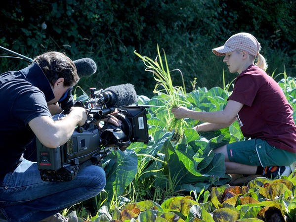 Ein Kameramann filmt eine Person, die in einem Gemüsefeld kniet und eine Pflanze begutachtet. Der Kameramann ist in der Hocke und hält eine professionelle Videokamera mit einem großen Mikrofon.
