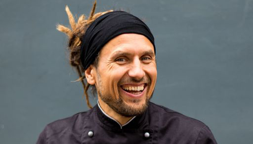 Ein Porträt eines lachenden Mannes mit Ziegenbart und blonden Dreadlocks, die von einer schwarzen Mütze bedeckt sind. Er trägt eine schwarze Kochjacke und schaut fröhlich zur Seite. Der Hintergrund ist eine graue Wand.