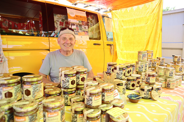 Eine ältere Person sitzt vor einem gelben Fahrzeug und einer Auslage mit vielen Gläsern oder Dosen, die dicht nebeneinander aufgestellt sind.
