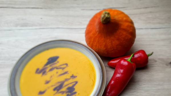 Eine Schale mit cremiger Kürbissuppe auf einem hellen Tisch, daneben ein kleiner dekorativer Kürbis und zwei rote Chilischoten. Die Suppe ist leuchtend orange und wirkt herbstlich-wärmend.