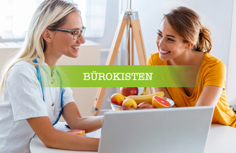 Zwei Frauen sitzen an einem Tisch, eine davon mit Laptop, und sprechen miteinander, daneben eine Schale mit Obst. Darüber liegt ein grünes Banner mit der Aufschrift 'BÜROKISTEN'.