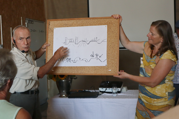 Zwei Personen, ein Mann und eine Frau, stehen vor einer weißen Tafel. Der Mann hält einen Stift und die Frau zeigt auf die Tafel, auf der Schriftzeichen zu sehen sind.
