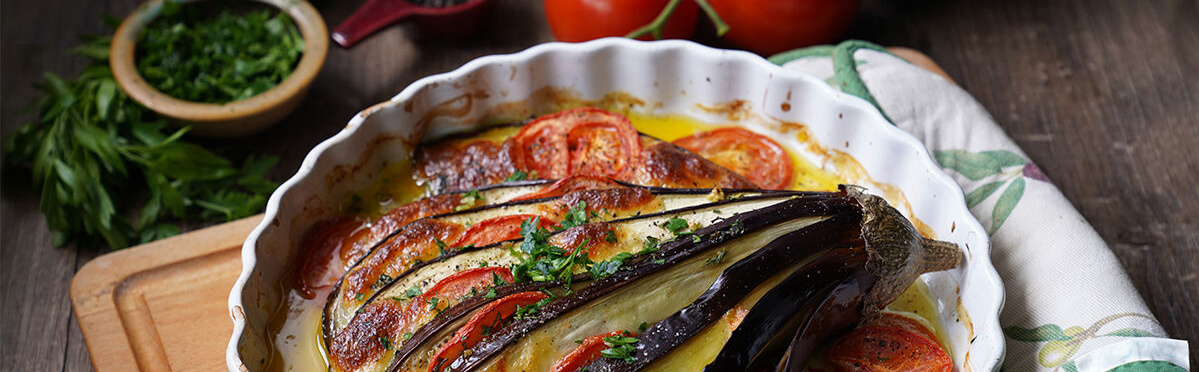 Ein bunt geschichtetes Auberginen-Tomaten-Gratin in einer weißen Auflaufform. Die Aubergine ist fächerförmig eingeschnitten und mit Tomaten- und Kartoffelscheiben gefüllt. Serviert auf einem rustikalen Holztisch, garniert mit frischen Kräutern.