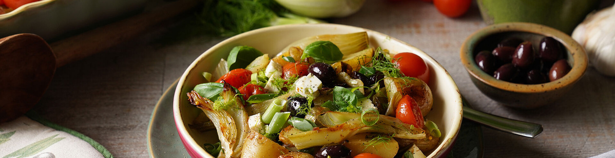 Ein bunter Salat mit gebratenem Fenchel, Cherrytomaten, Oliven, Frühlingszwiebeln und frischen Kräutern in einer Schale. Daneben eine kleine Schale mit schwarzen Oliven. Die Szene ist hell und einladend gestaltet.