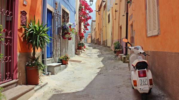 Eine charmante, schmale Gasse mit bunt gestrichenen Häusern, Blumentöpfen und Wäscheleinen. Einladendes mediterranes Flair – vermutlich in Südeuropa.
