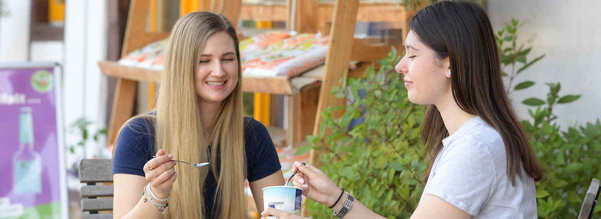 Zwei Frauen sitzen draußen an einem Tisch und essen gemeinsam Eis. Auf dem Tisch steht ein kleiner Korb mit Sonnenblumen.