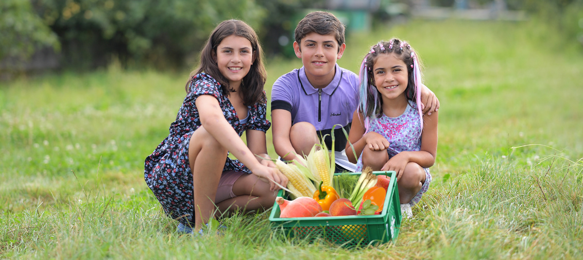 Drei Kinder sitzen auf einer Wiese neben einer grünen Kiste voller frischem Gemüse und Obst, darunter Mais, Paprika und Melonen. Alle lächeln in die Kamera.