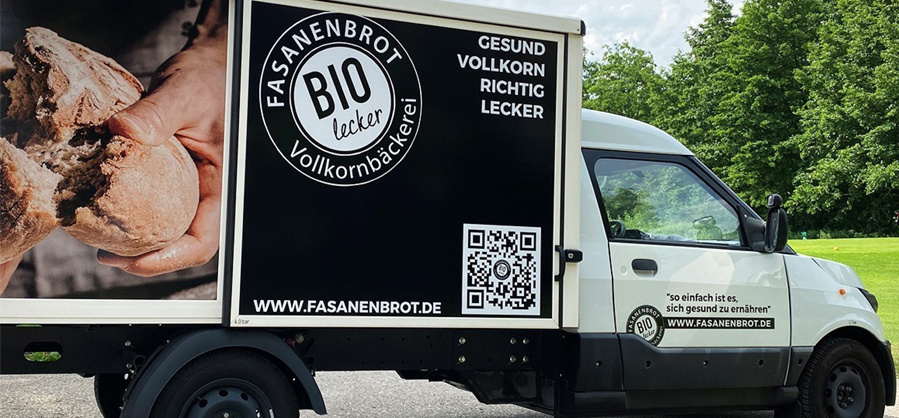 Ein weißer Lieferwagen mit einer schwarzen Werbefläche auf der Seite. Auf der schwarzen Fläche steht in großen Buchstaben "FASANENBROT BIO lecker Vollkornbäckerei" mit einem Logo. Darunter befindet sich ein QR-Code und die Website-Adresse "WWW.FASANENBROT.DE". Rechts daneben steht "GESUND VOLLKORN RICHTIG LECKER". Auf der Fahrertür des Lieferwagens steht der Slogan "so einfach ist es, sich gesund zu ernähren" mit einem "BIO"-Zeichen und erneut der Website-Adresse. Ein Teil der Seitenwand zeigt ein Bild von Händen, die frisches Brot halten. Im Hintergrund sind grüne Bäume und ein blauer Himmel zu sehen.