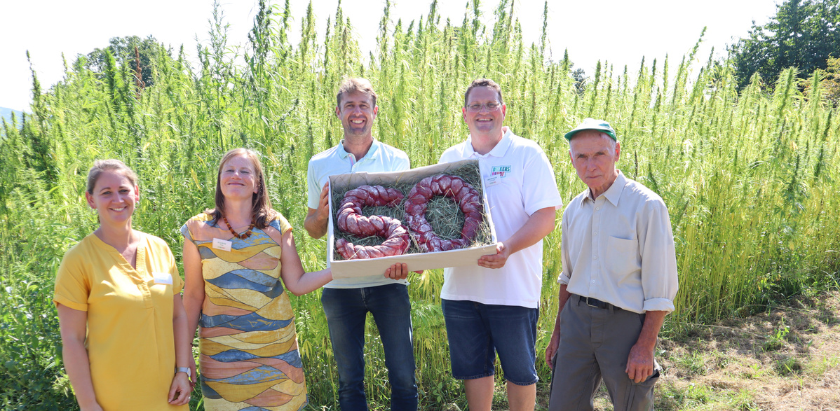 Eine Gruppe von fünf Erwachsenen steht auf einem Feld mit hohen grünen Pflanzen. Die beiden Personen in der Mitte halten gemeinsam eine große Holzkiste, in der sich ein Brot in Form der Zahl '50' befindet, das ein 50-jähriges Jubiläum symbolisiert.