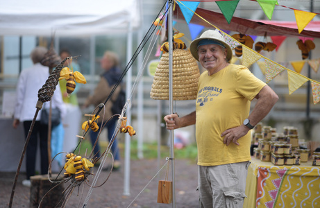 Eine Person in einem gelben Hemd und Hut lächelt, während sie neben dekorativen Sonnenblumen und Honiggläsern an einem Marktstand steht.