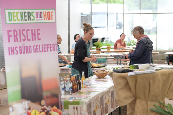 Ein Jungpflanzenstand mit einem Schild, das "DECKERS BIOHOF - FRISCHE INS BÜRO GELIEFERT" anzeigt. Im Vordergrund sind Tische mit verschiedenen Produkten und einige Personen zu sehen, die an den Ständen einkaufen.